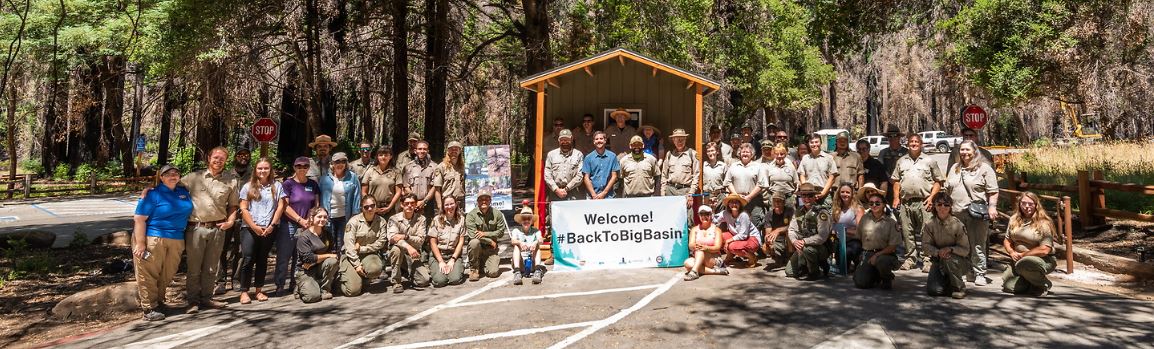 A large group of people are posing happily around a wooden entryway outline over a road. In the middle is a banner reading Welcome Back To Big Basin.